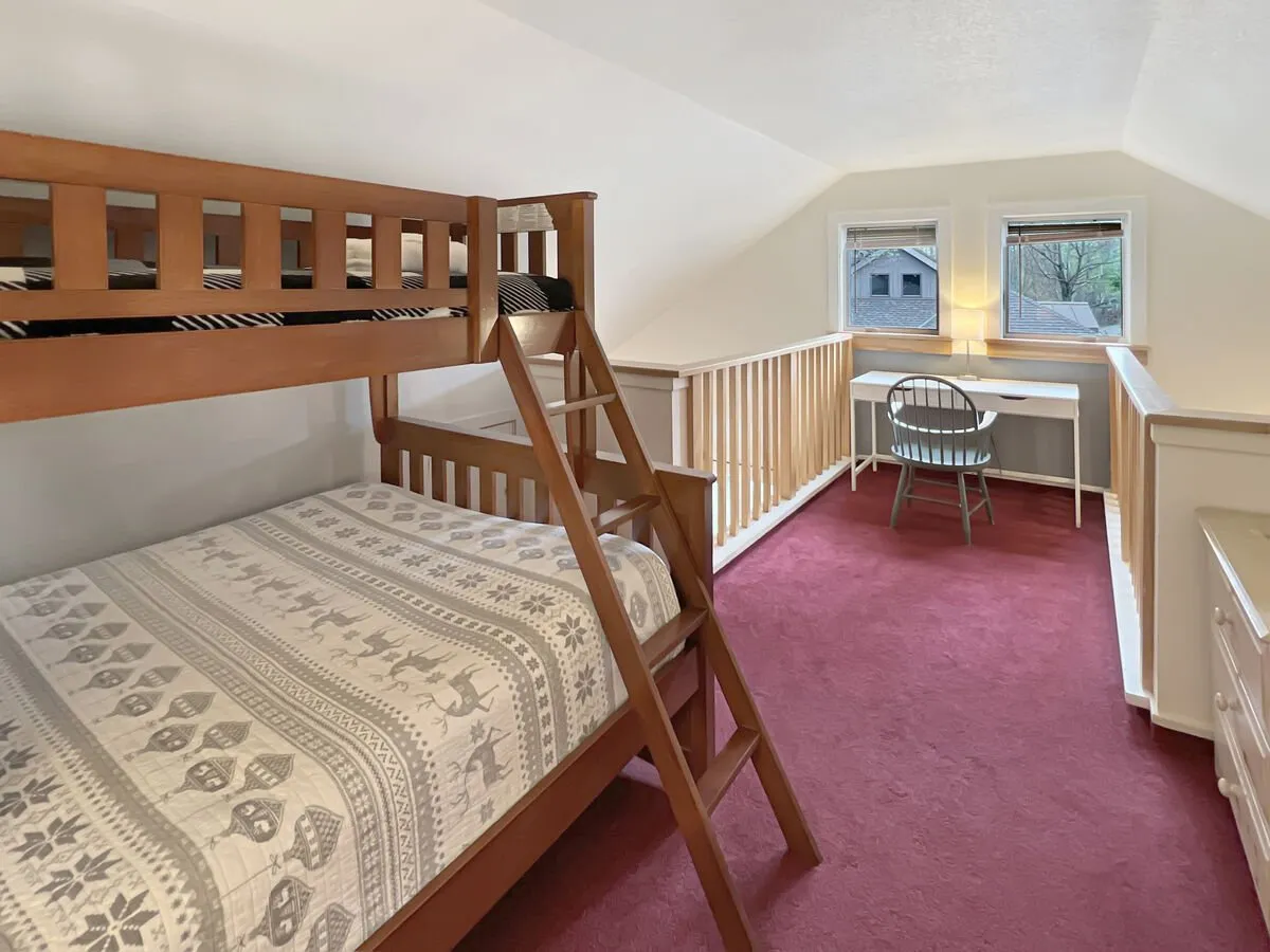 A loft space featuring a bunk bed setup with a queen bed below and a twin bed above. The room is brightened by windows that allow in natural light, and a study desk sits nearby, providing an inviting space for work or study. The playful, adaptable vibe is ideal for both relaxation and productivity.