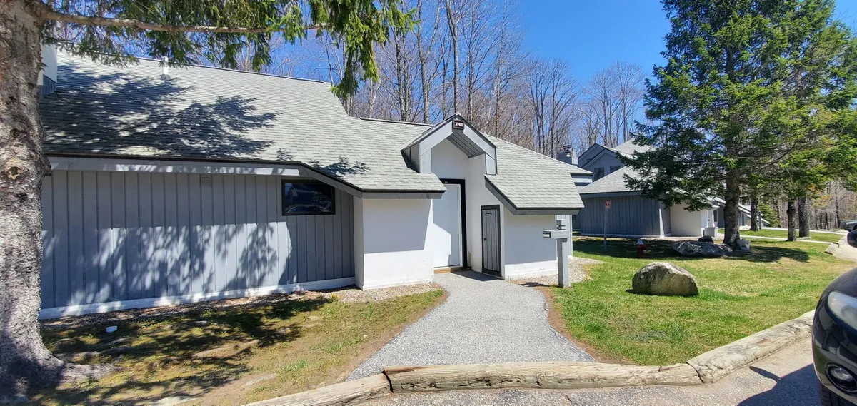 This exterior image features the front entrance of Woods Village 43, framed by a landscaped pathway and surrounded by mature trees. The inviting porch suggests a welcoming space for guests to gather, while the natural setting hints at outdoor adventures and relaxation.