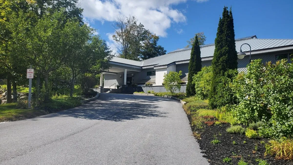 The image shows the sloped driveway leading to the main entrance, bordered by lush greenery and landscaped areas. The view emphasizes both the accessibility and curb appeal of the property, providing a sense of place and room for outdoor gatherings.