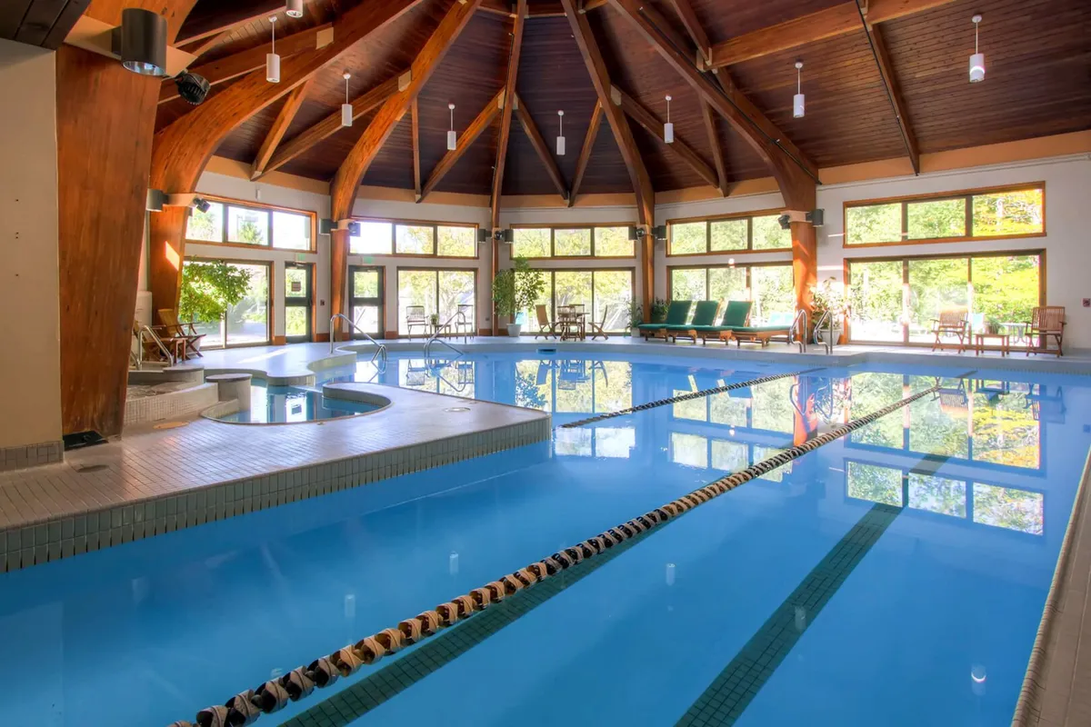 A modern indoor recreation space featuring a large swimming pool and an adjacent jacuzzi/hot tub. The area is framed by expansive windows and rustic wooden beams, with seating for relaxation and social gatherings, capturing a mood of calm and rejuvenation.