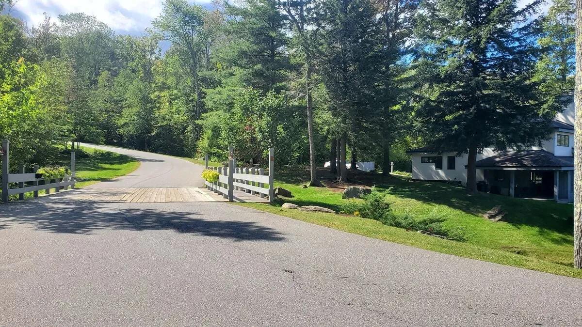 A tranquil, tree-lined driveway leads to a wooden bridge and lush greenery, with a house visible in the background. The landscape invites walking, biking, and outdoor exploration, perfectly suited to families and adventurers alike.