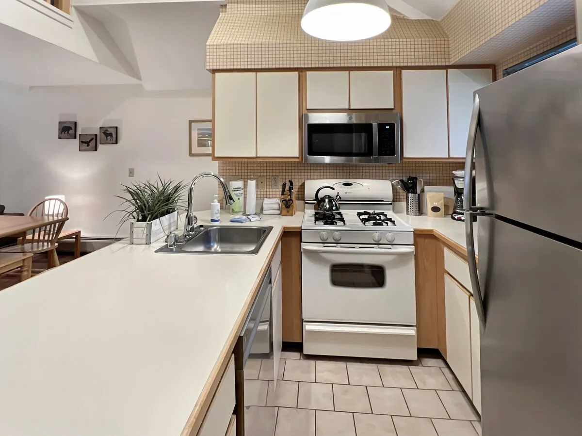 A kitchen island with stainless steel appliances set beside a sun-drenched dining area, flooded with natural light from large windows. The scene captures the heart of meal preparation, family gatherings, and sociable moments, all wrapped in a warm and welcoming mood.