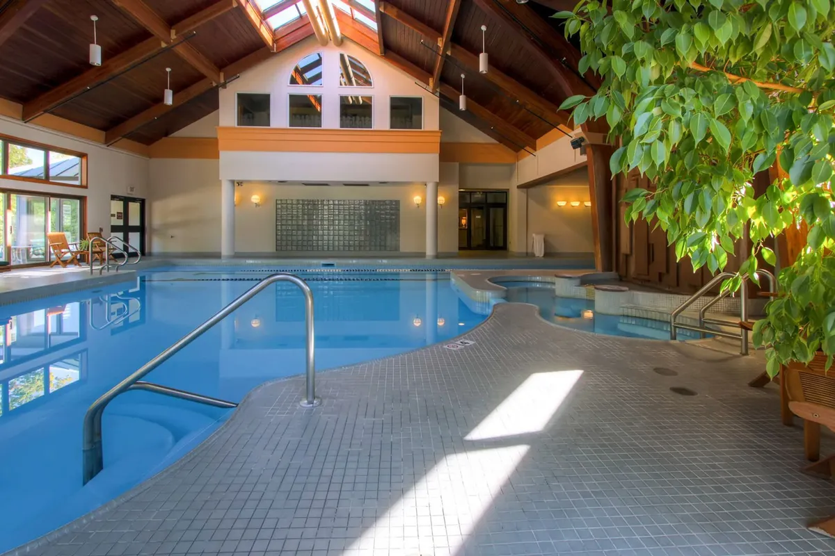 A serene indoor swimming pool surrounded by large windows with views of lush greenery. Exposed wooden beams and natural sunlight filtering through skylights create a warm, inviting atmosphere. Guests can be seen swimming, relaxing by the poolside, and socializing with friends, highlighting the communal and rejuvenating aspects of the amenity.