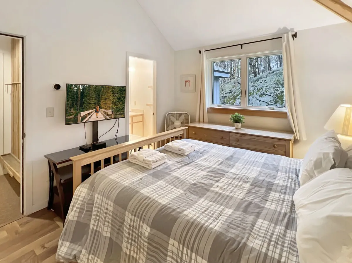 A tranquil bedroom with a king-sized bed, a television situated across from the bed, a dresser, and a window offering a view outside. The space is calm and uncluttered, providing a restful environment ideal for unwinding after a day of activities.
