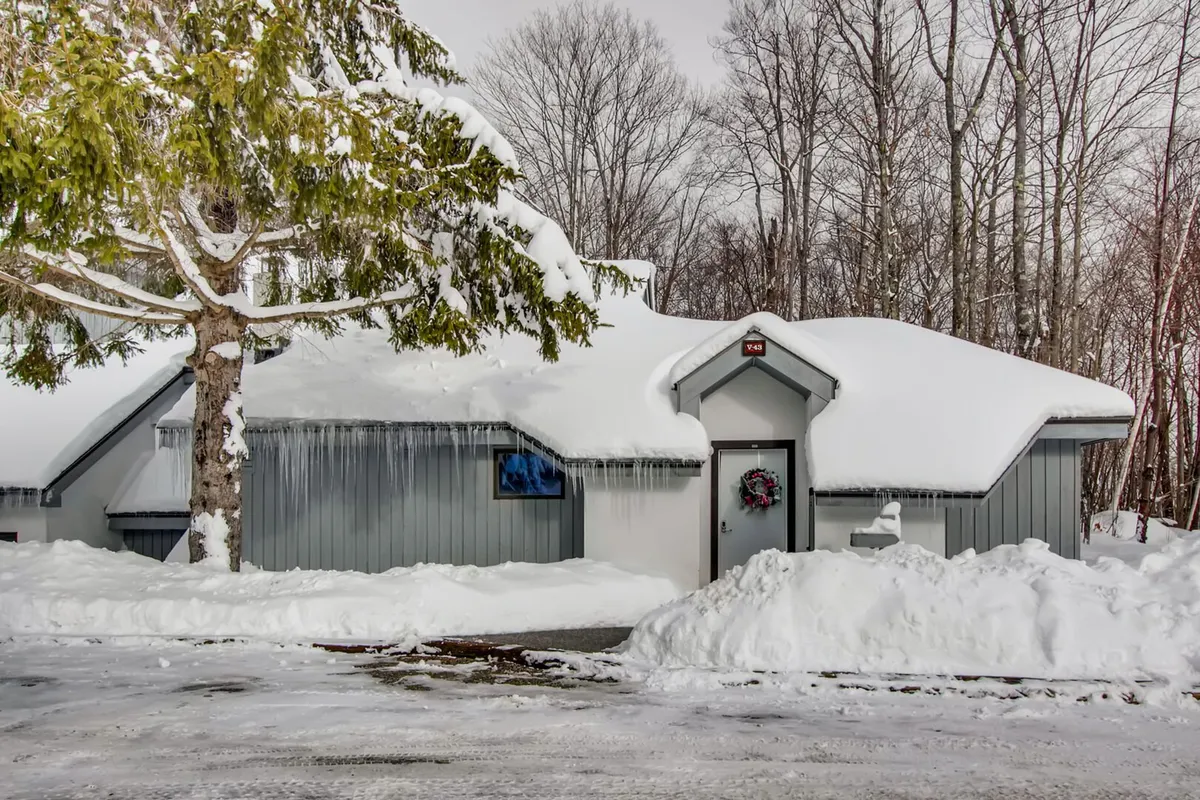 A snow-covered roof and decorative wreath highlight a peaceful home nestled among tall, snow-dusted trees. The inviting exterior suggests cozy gatherings and wintertime fun after days spent outdoors.