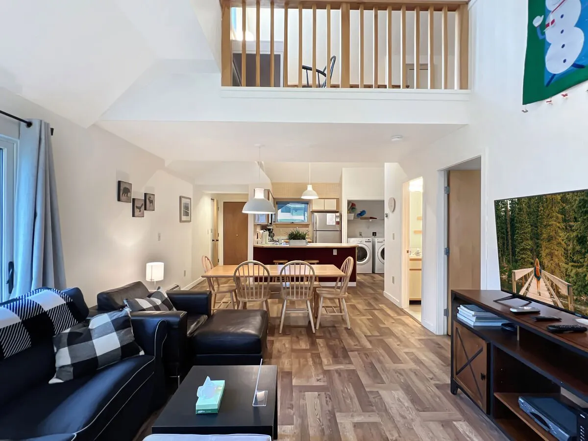 An open-plan living space with black leather sofas, a dining table with wooden chairs, a sleek open kitchen equipped with modern appliances, and a glimpse of a balcony railing letting in natural light. The area looks perfect for family gatherings, casual dining, and relaxing evenings after outdoor adventures.