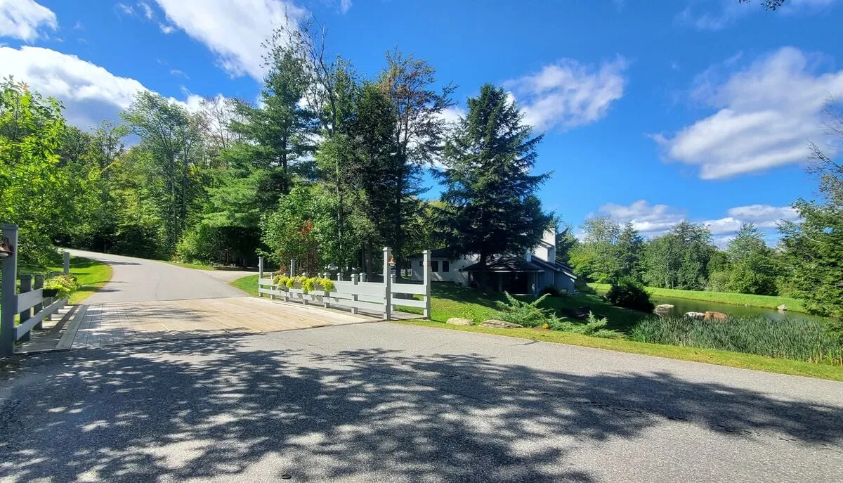 A tranquil exterior scene showcasing the house, entrance gate, mature trees, and a peaceful pond. The space feels open and invites guests to explore the natural surroundings, echoing the sense of discovery and relaxation after a day out.